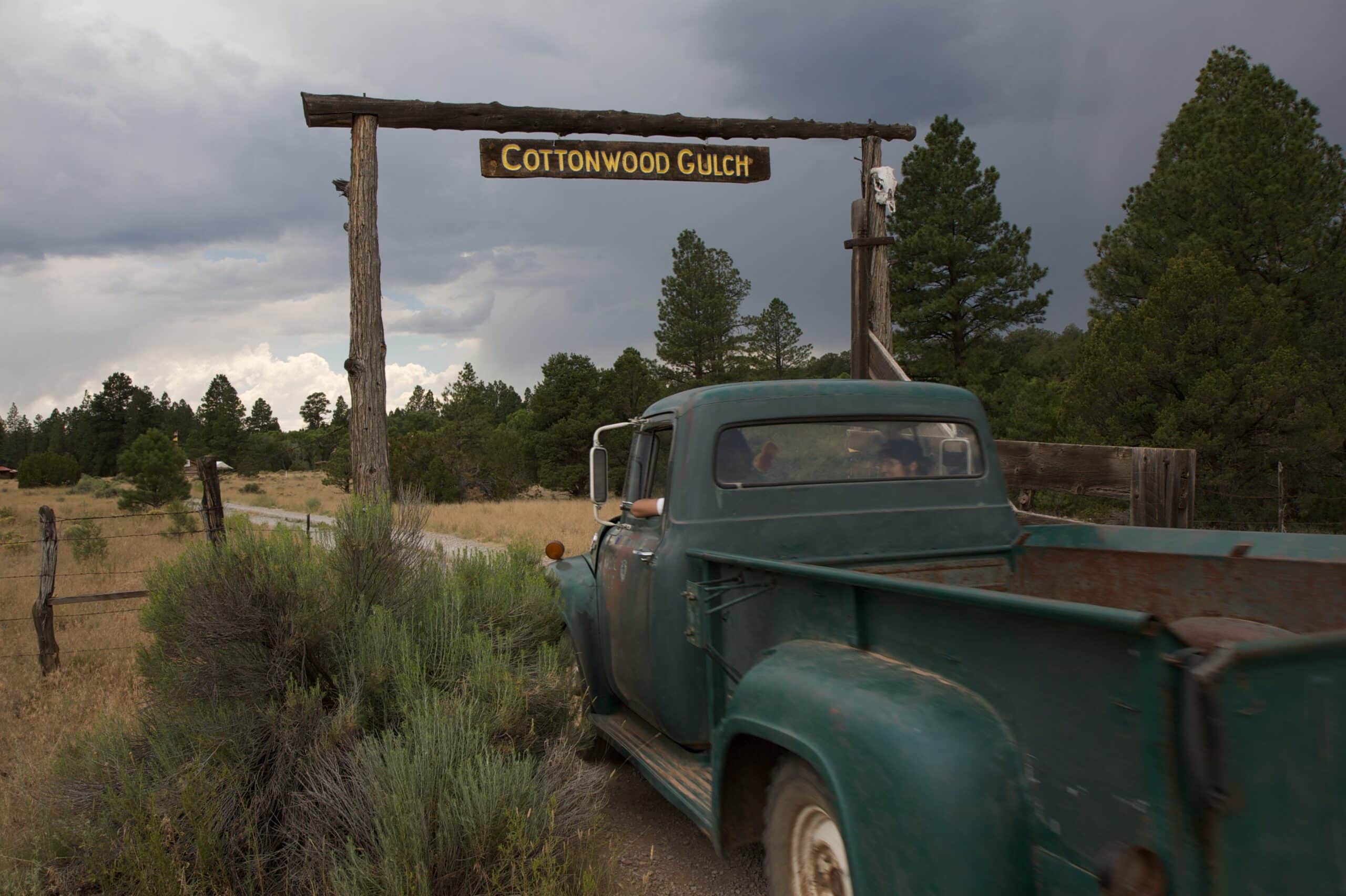 Old green pickup truck driving under wooden Cottonwood Gulch entrance sign on a dirt road surrounded by pine trees and cloudy sky