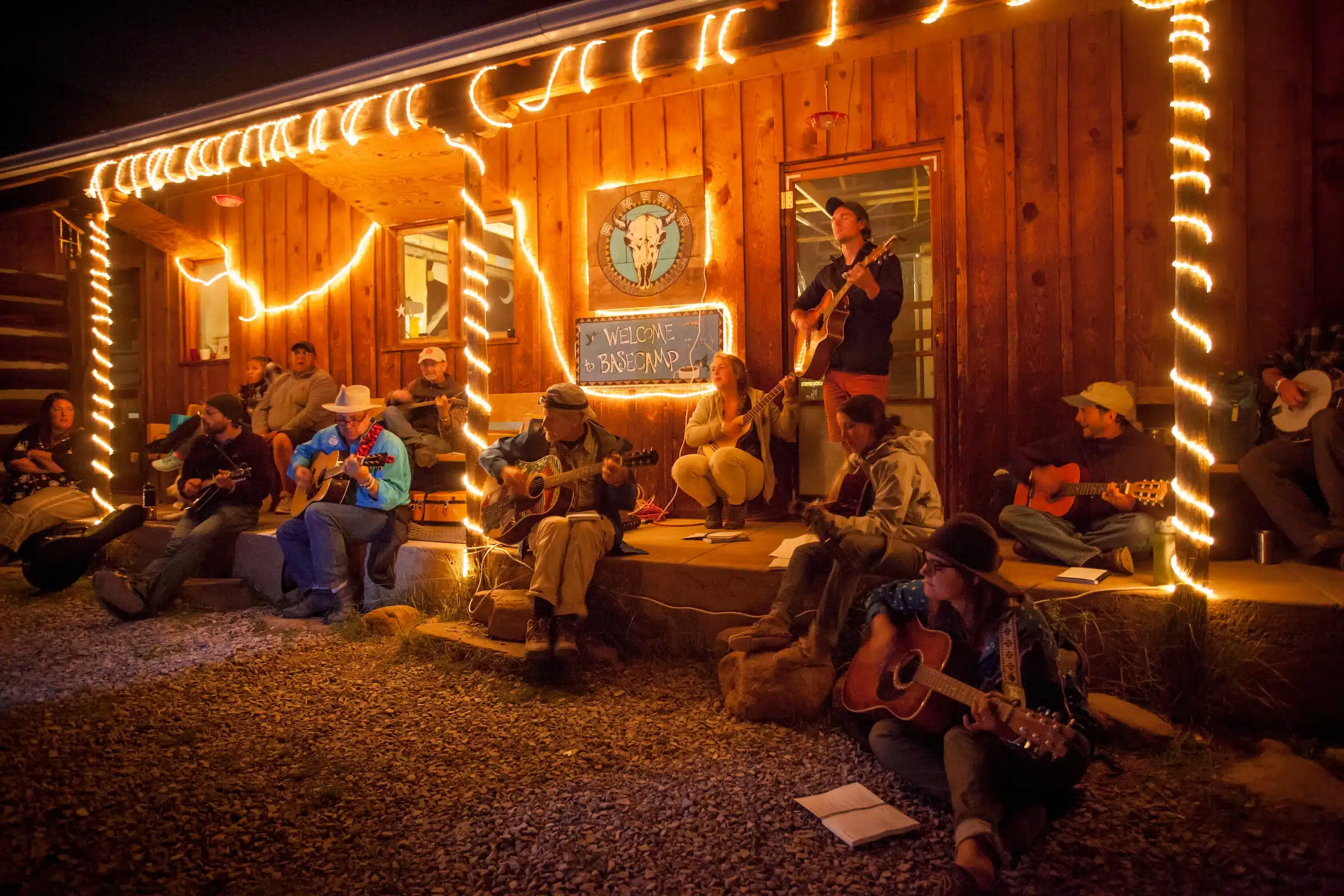 Musicians play guitars during an evening porch jam session outside a rustic wooden cabin lit with string lights and a ‘Welcome to Basecamp’ sign