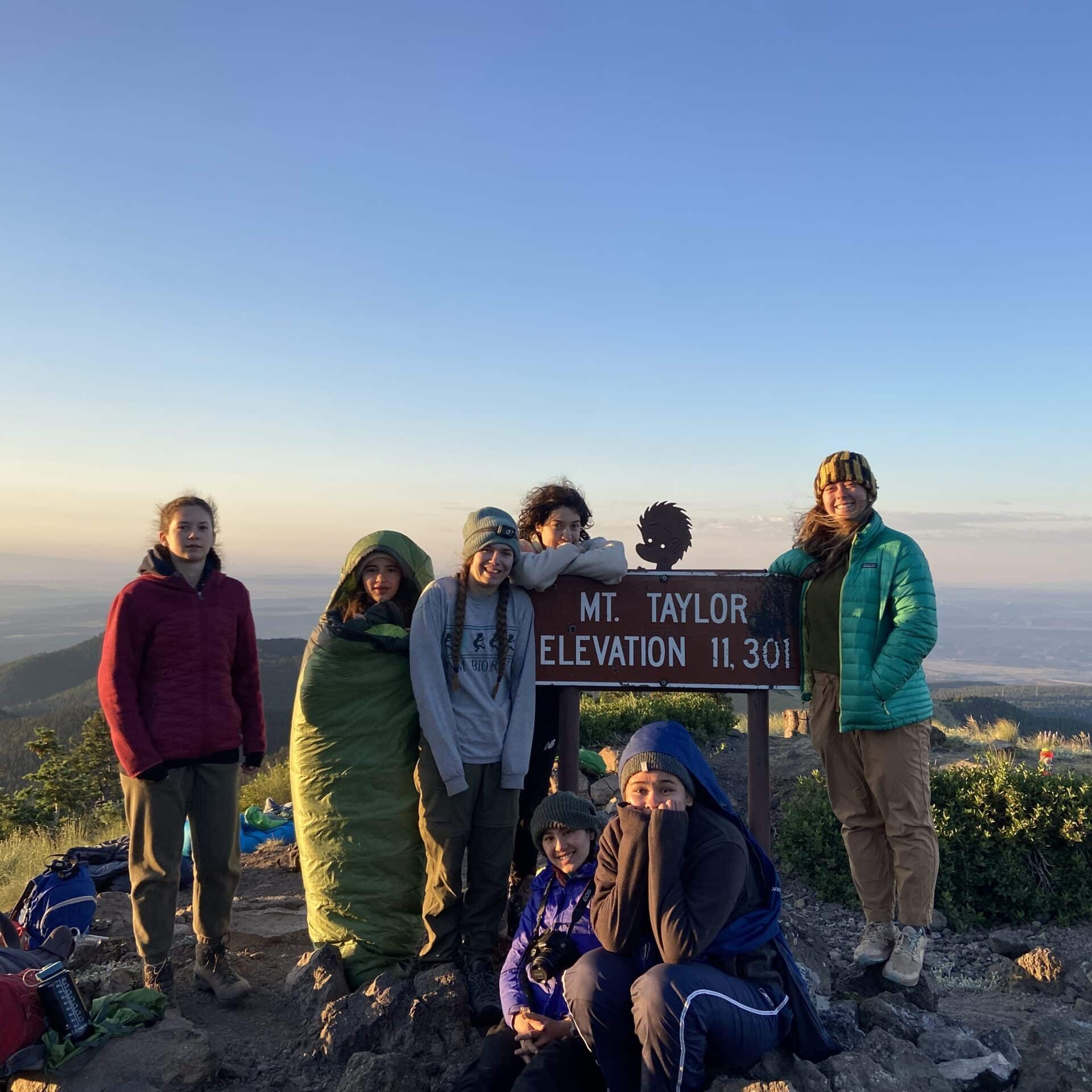 a group of girls in parkas and sleeping bags perch atop Mt Taylor at sunrise.